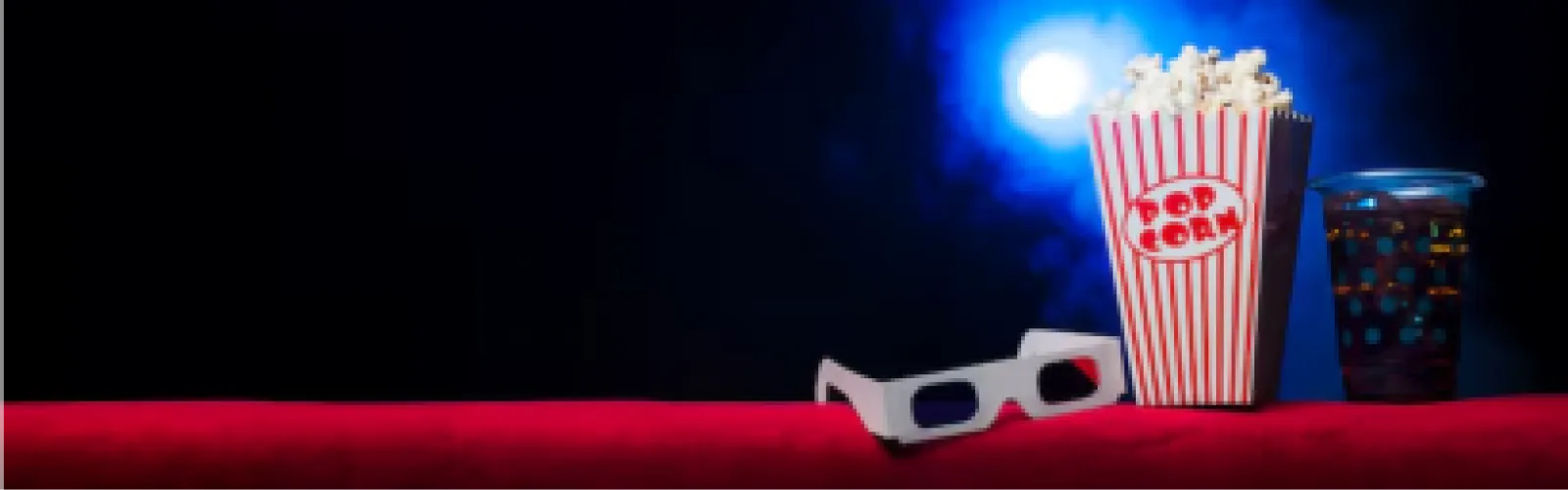 Red and white popcorn bucket filled with popcorn, 3-D glasses, and a cup of soda sitting on a red surface with black background